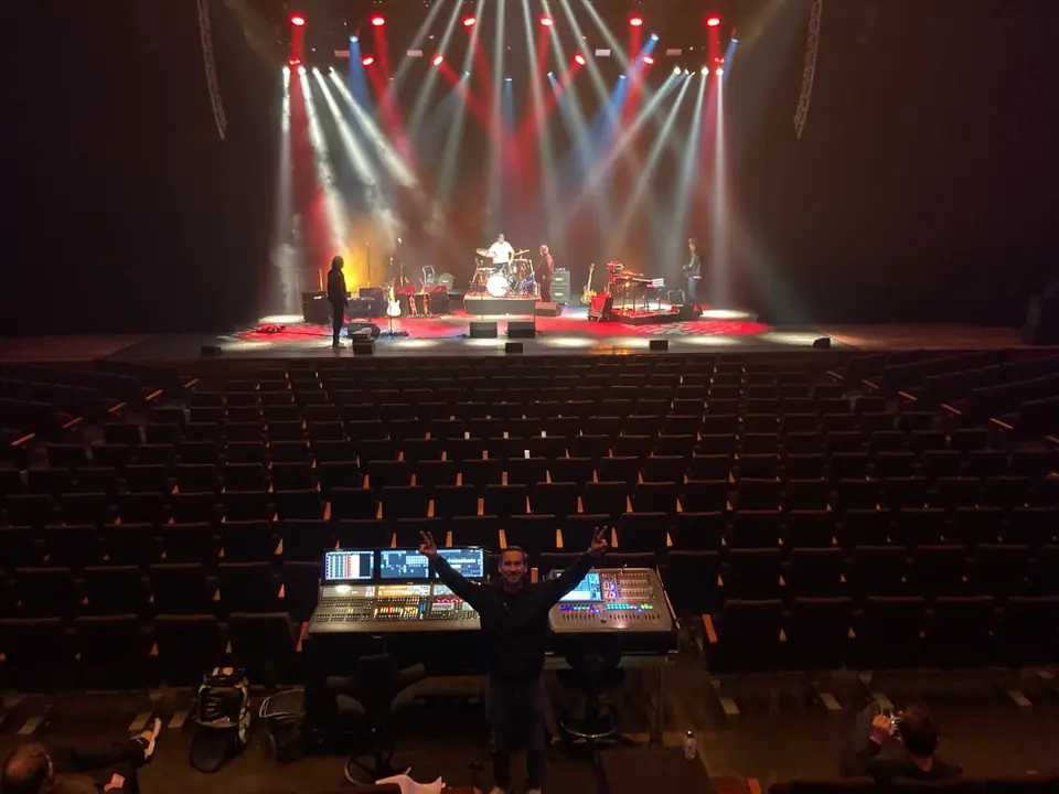 Paul Heijkens juicht met beide armen omhoog achter de lichttafel in een lege theaterzaal, op het podium rode en witte beams