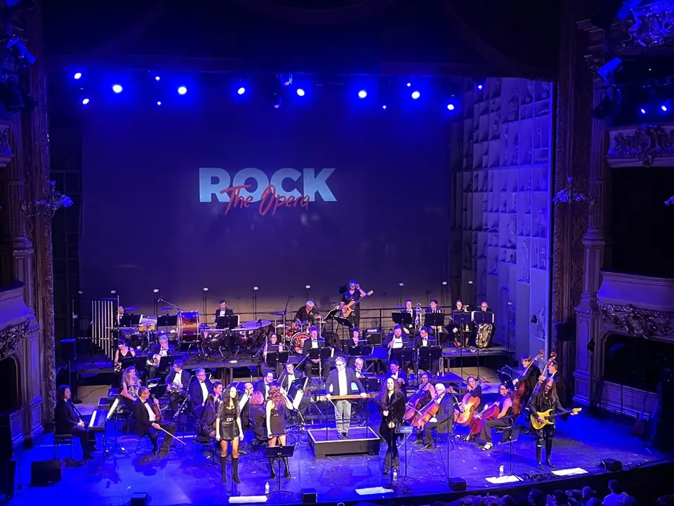 Rock the Opera: symfonieorkest, zangeressen en gitarist in blauw licht, het logo op het projectiedoek erachter