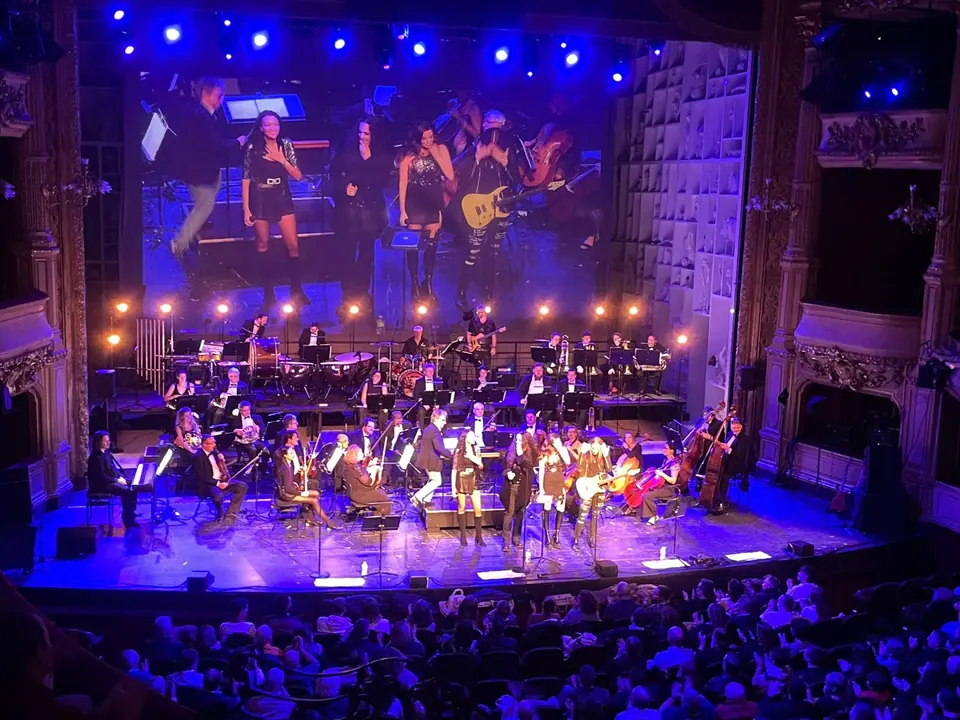 Rock the Opera: zangeressen en gitarist vooraan, orkest in blauw en warm licht, close-up op het doek