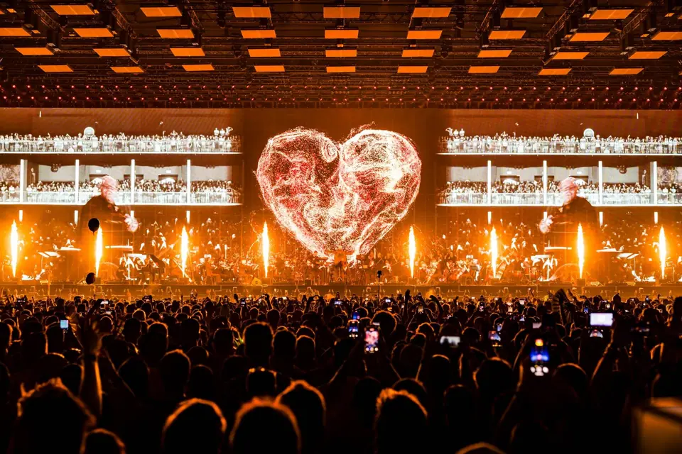 Tomorrowland 2025: orkest op het podium voor een hart van licht, met vuurfonteinen en oranje plafondschermen